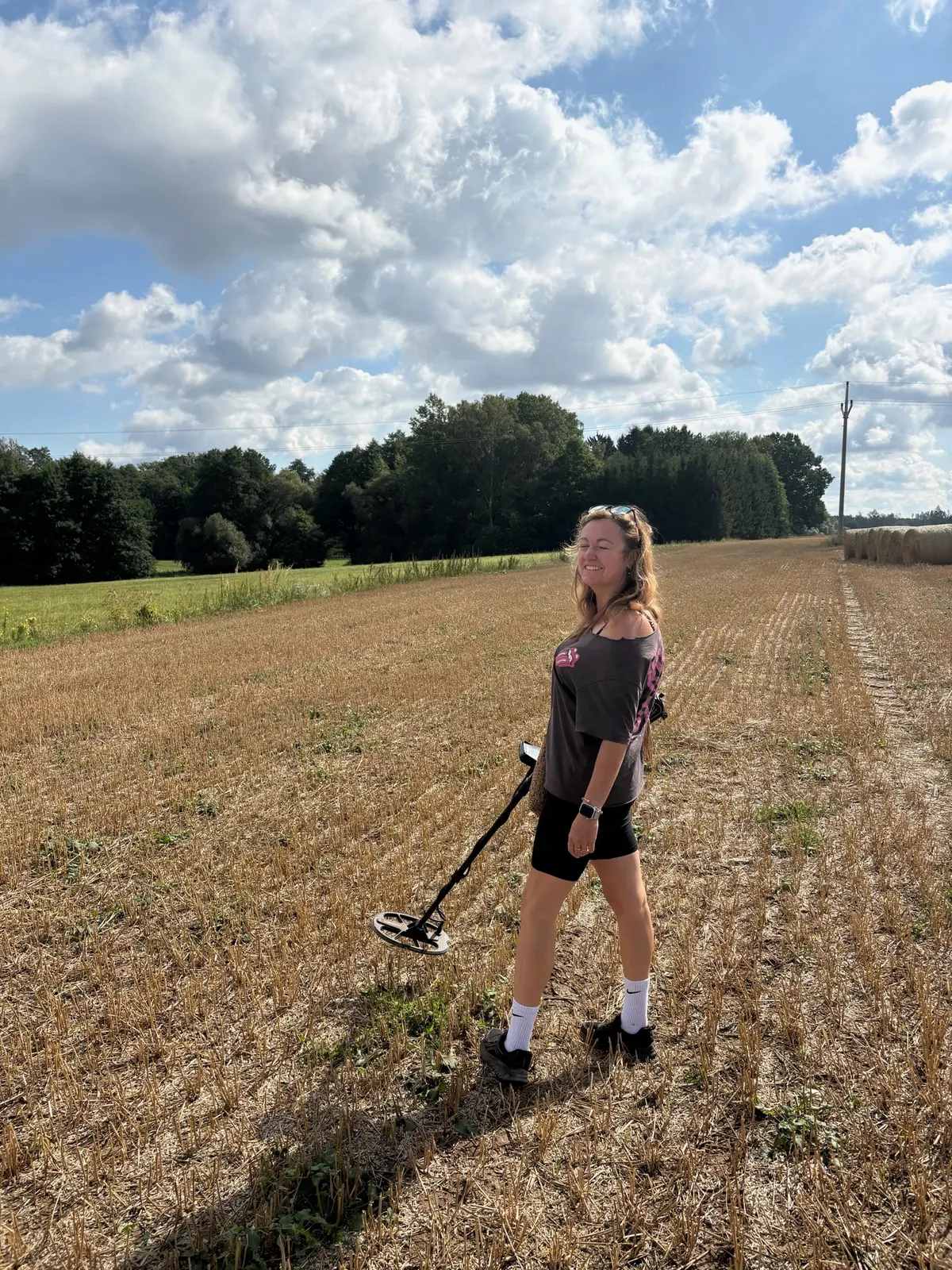 Žena s detektorem stojí na strništi, v pozadí baly slámy.