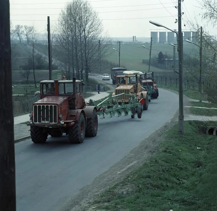 Kolona traktorů s nářadím na silnici, v čele červený kloubový traktor