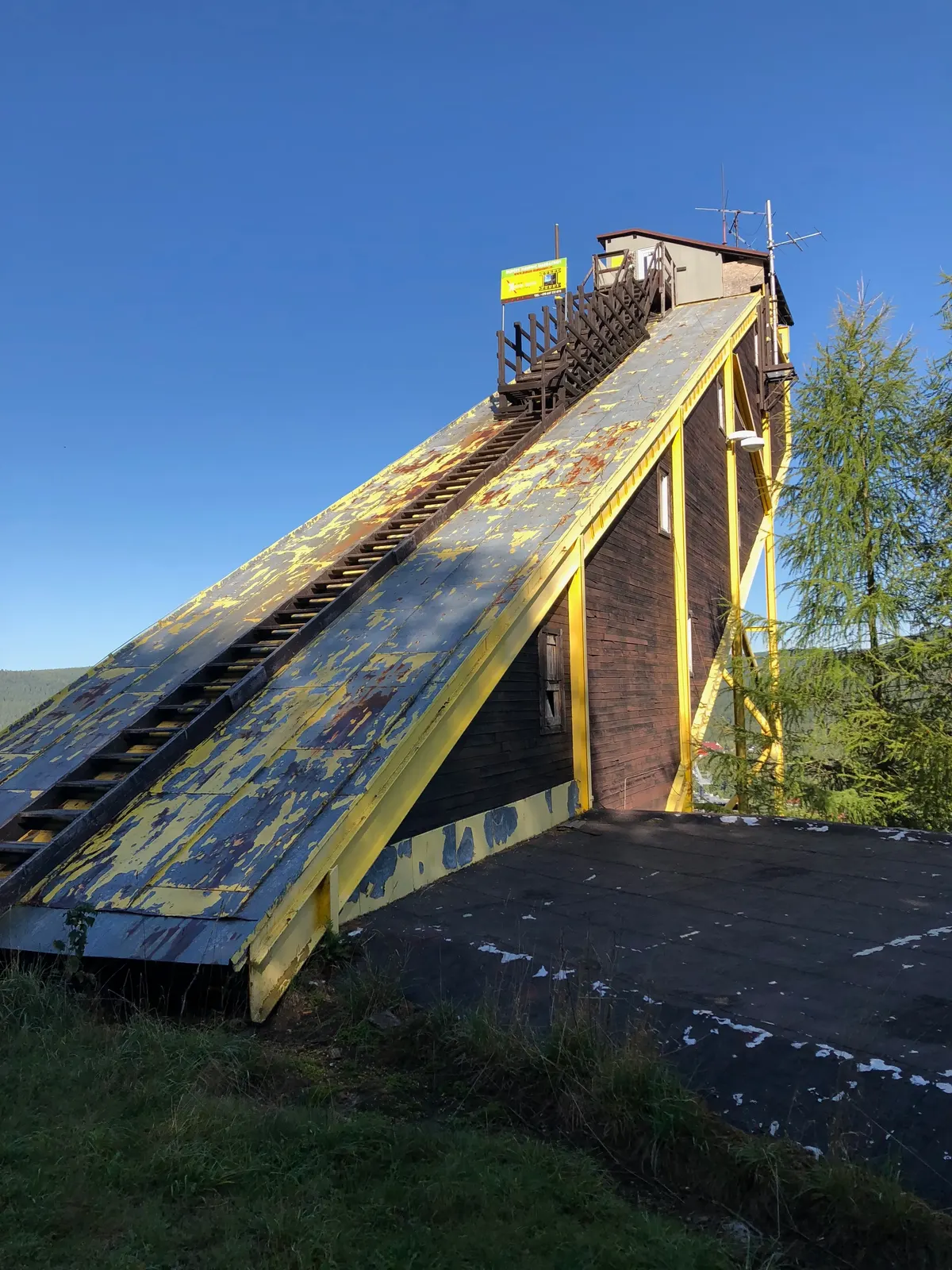 Dřevěná stavba u doskočiště se žlutou šikmou plochou a schodištěm, nátěr se loupe.