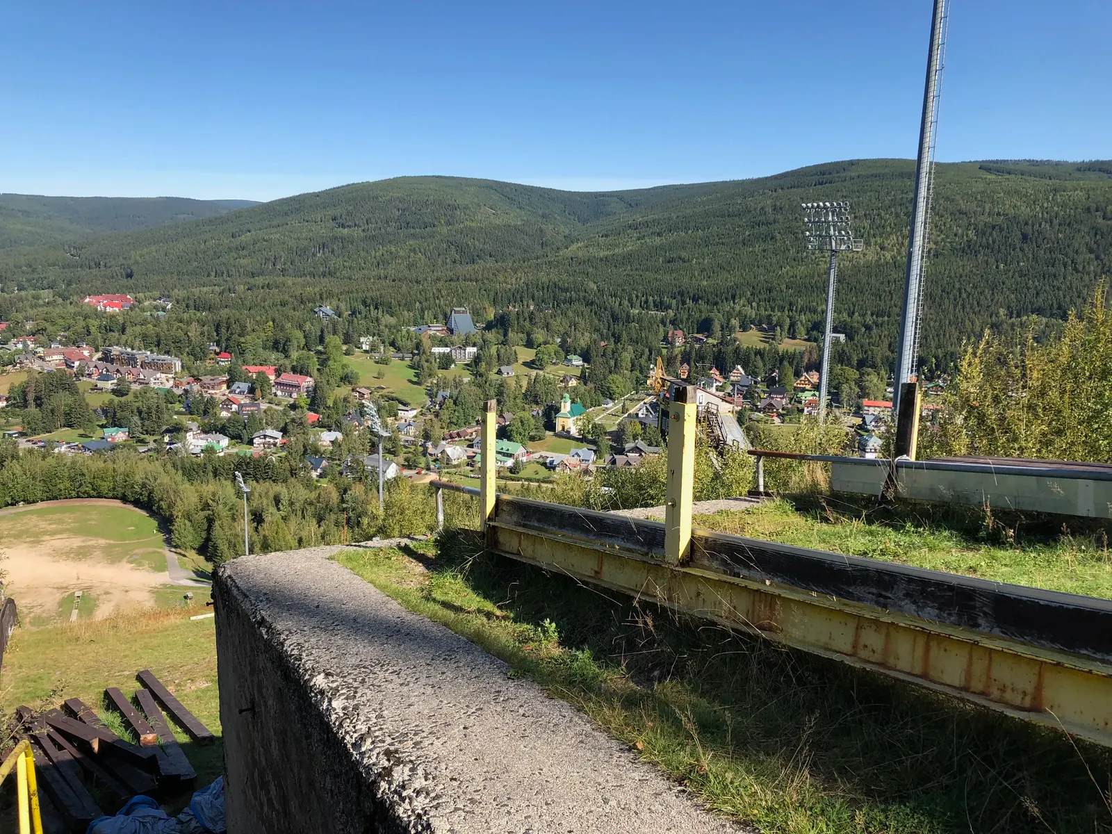 Betonová zídka a žluté sloupky, za nimi Harrachov a stožár osvětlení.
