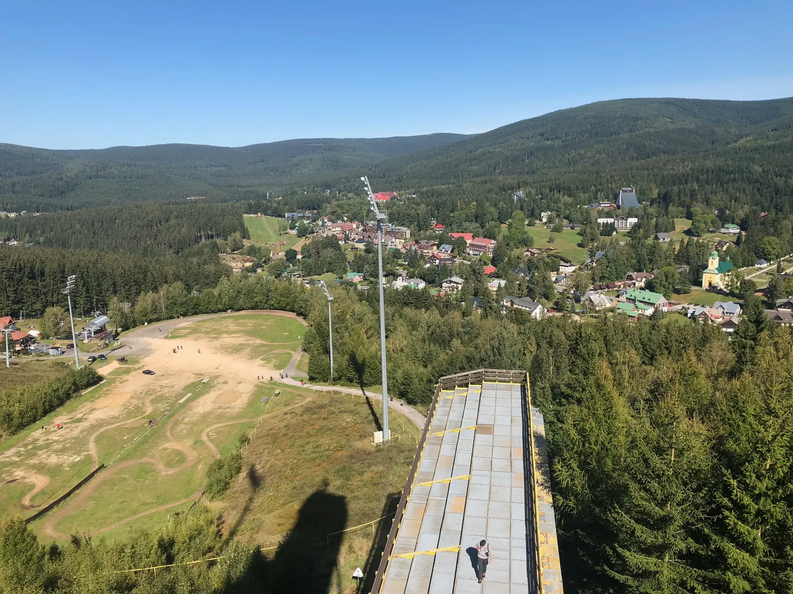 Tribuna shora, za ní výběh se stožárem osvětlení a Harrachov.