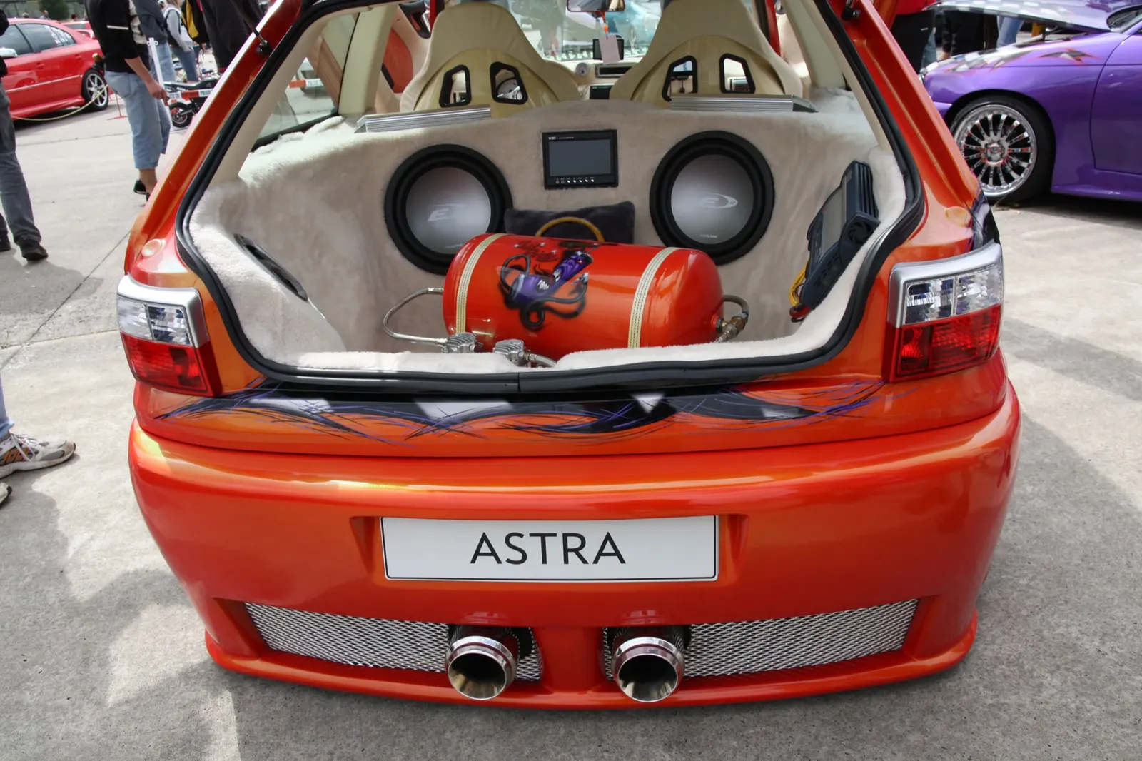 Oranžový Opel Astra s otevřeným kufrem, uvnitř dva subwoofery, air tank a monitor