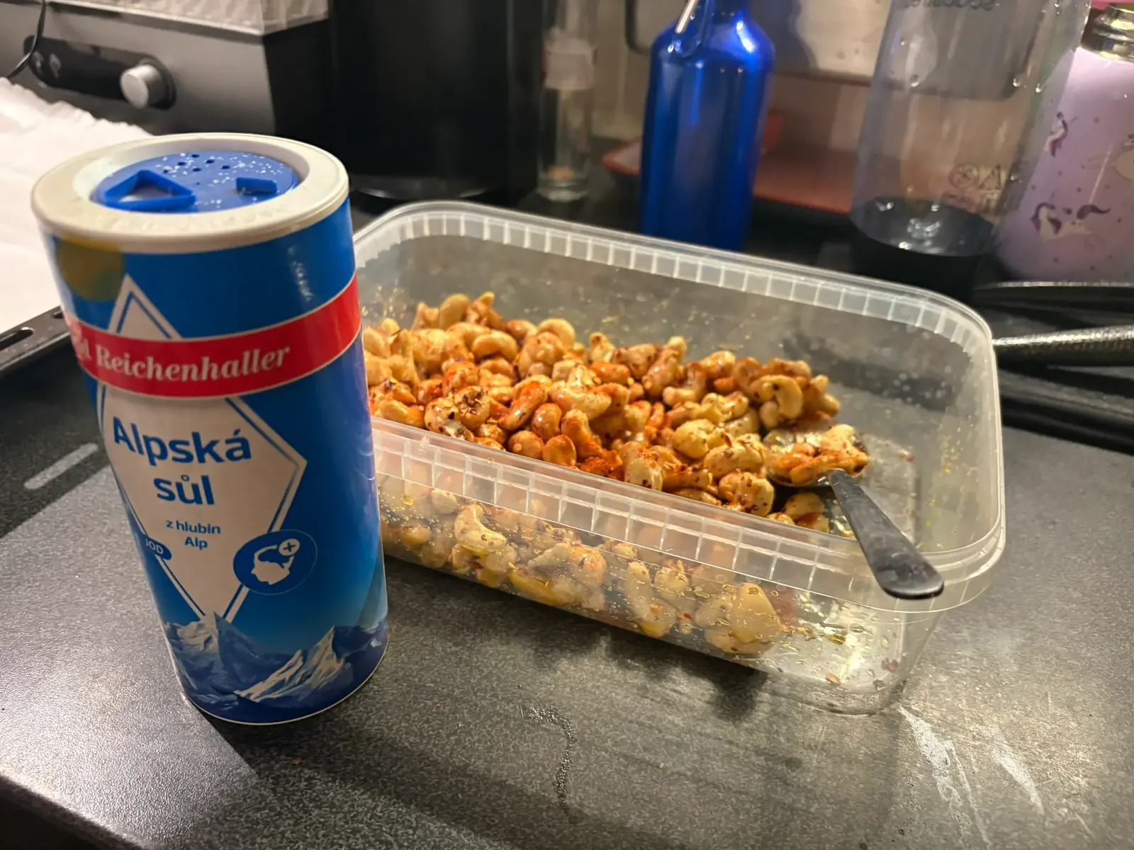 Kešu ořechy obalené medem, paprikou a chilli v plastové misce se lžící, vedle sůl