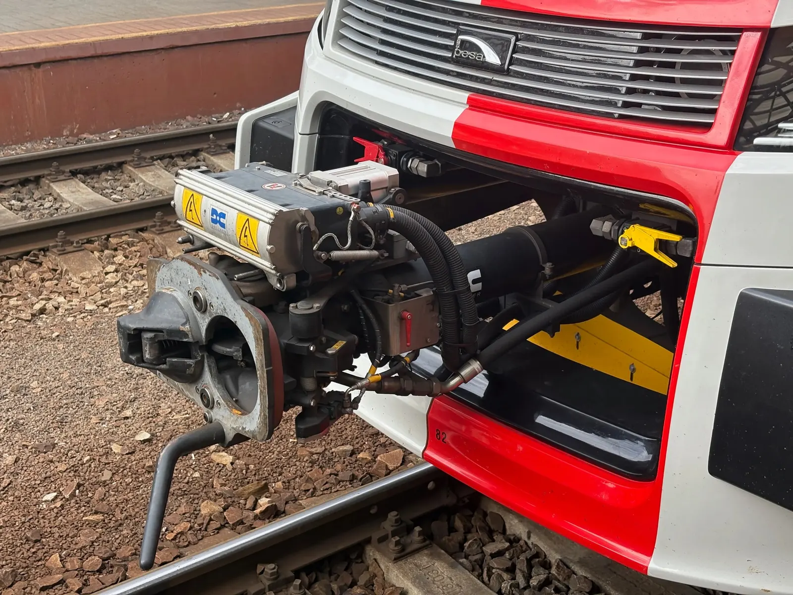Detail automatického čelního spřáhla RegioFoxu vyklopeného před vlak. Kovová hlava s mechanickými čelistmi vpředu, nad ní vodorovná elektrická krabička značky Dellner se žlutou samolepkou o vysokém napětí. Vlevo pryžové hadice pneumatiky, vpravo kabelové svazky.