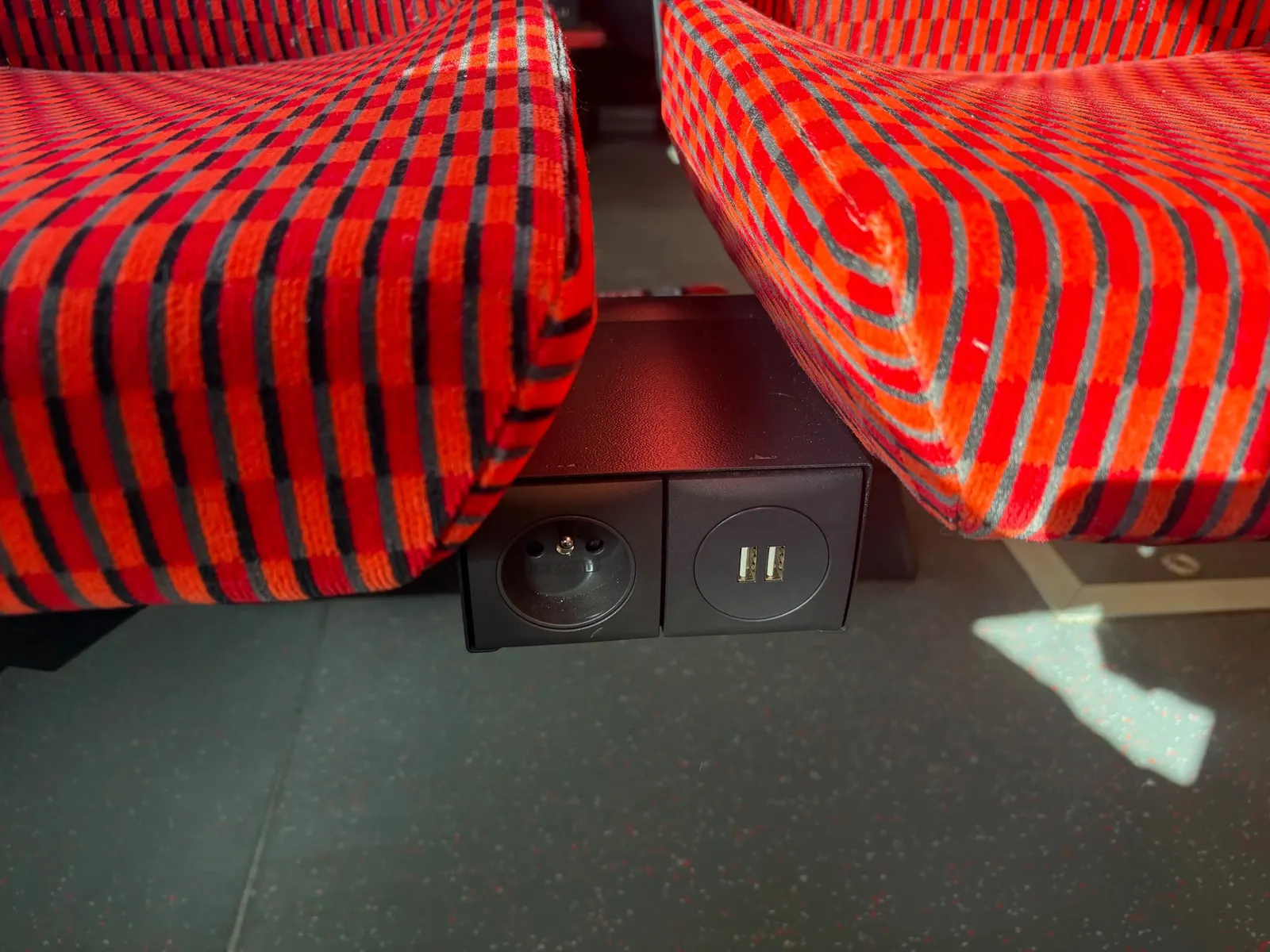 Detail napájecího modulu mezi dvěma sedačkami. Vlevo česká zásuvka 230 V, vpravo dvě USB-A porty. Modul je namontovaný na svislém středovém sloupku pod sedačkami.