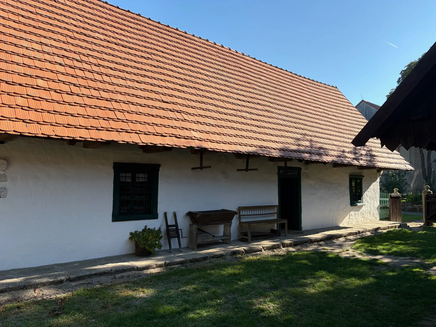 Dlouhá bílá chalupa s červenou taškovou střechou, plotem před domem, lavičkou a malým stolkem u vchodu
