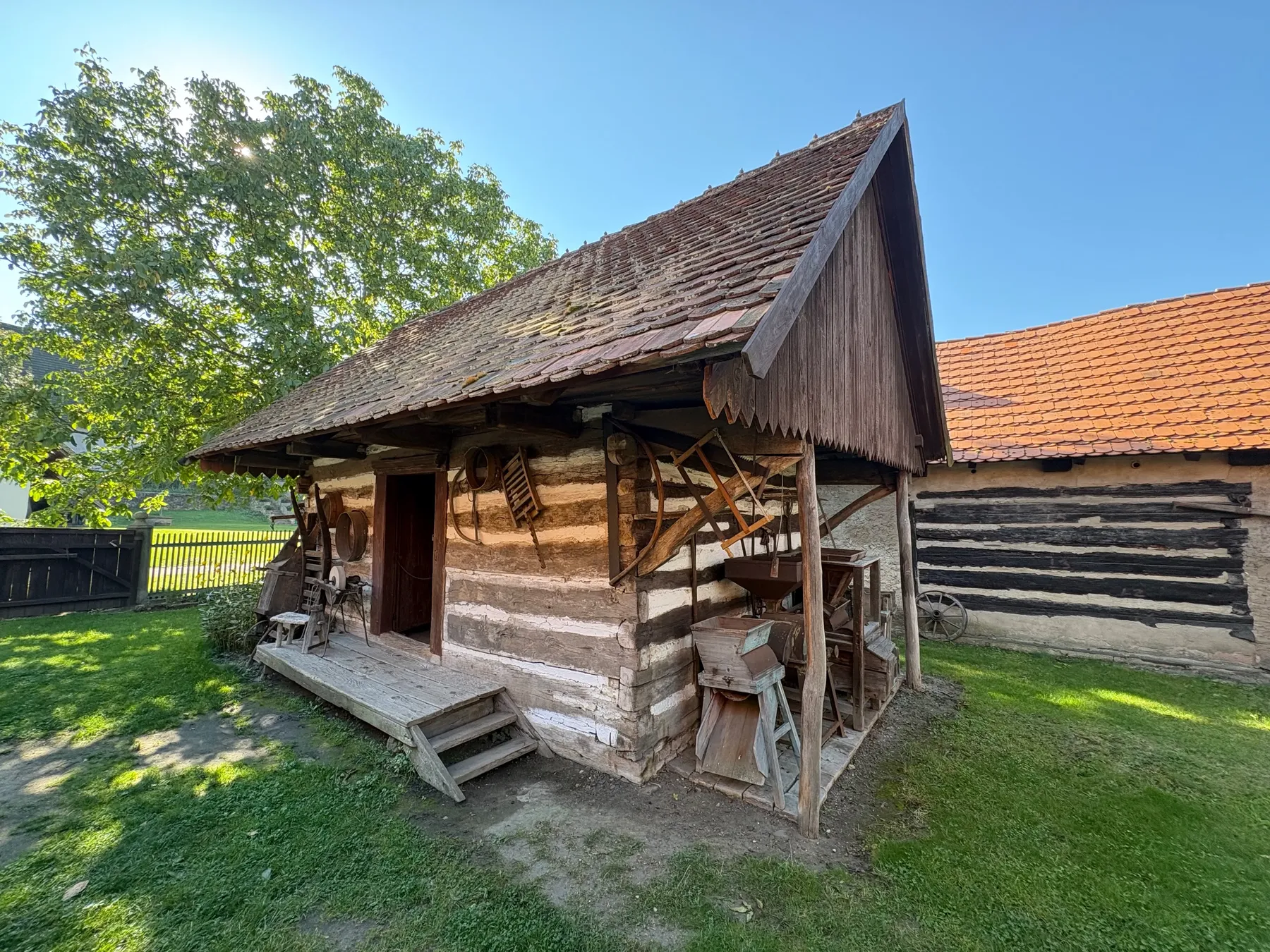 Malý dřevěný roubený špýchar s červenou taškovou střechou a bedněným štítem, sluneěná lavička