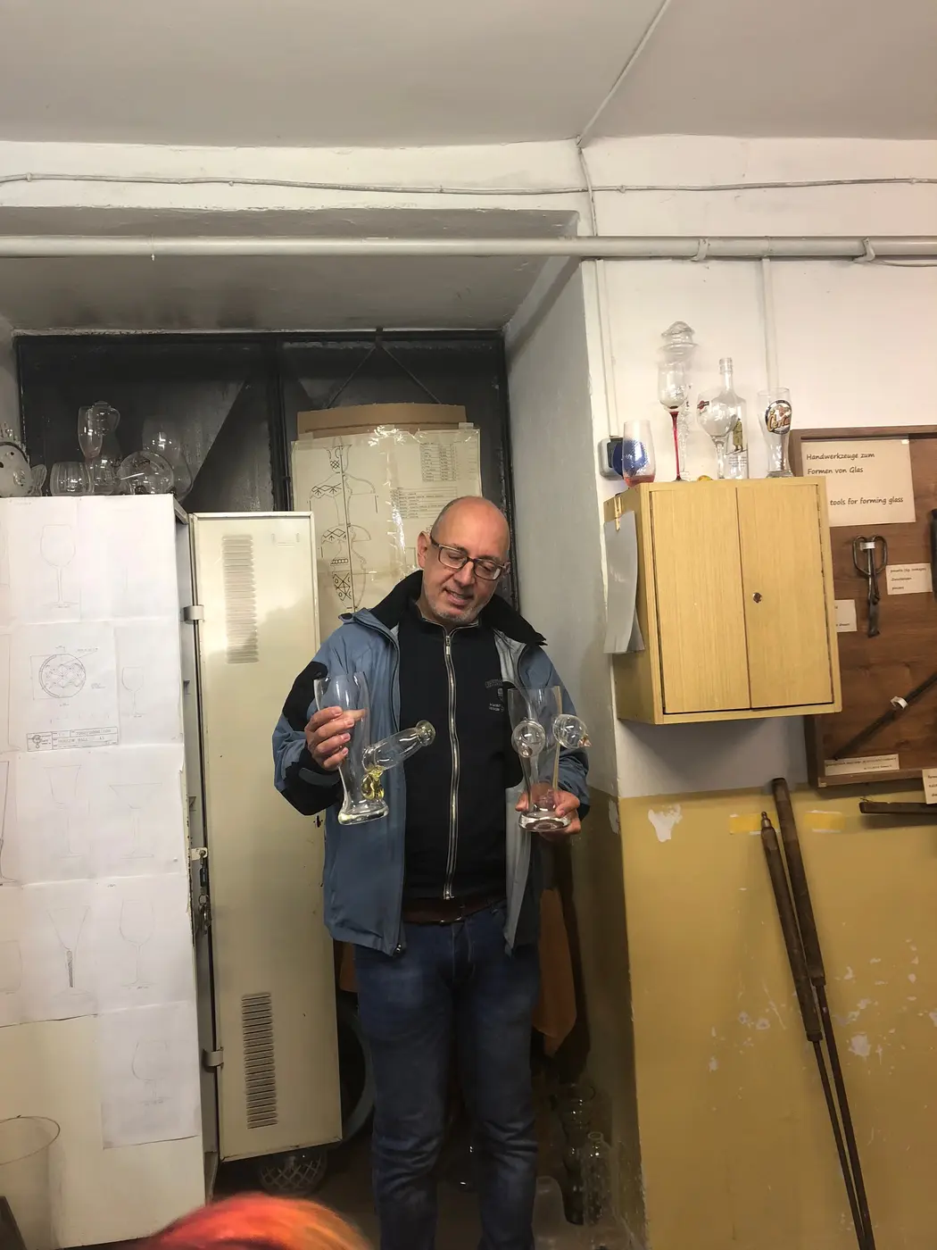 Průvodce v modré bundě drží tři sklenice různých tvarů, vlevo skříňka s papírovými výkresy tvarů, vpravo vitrína s nástroji