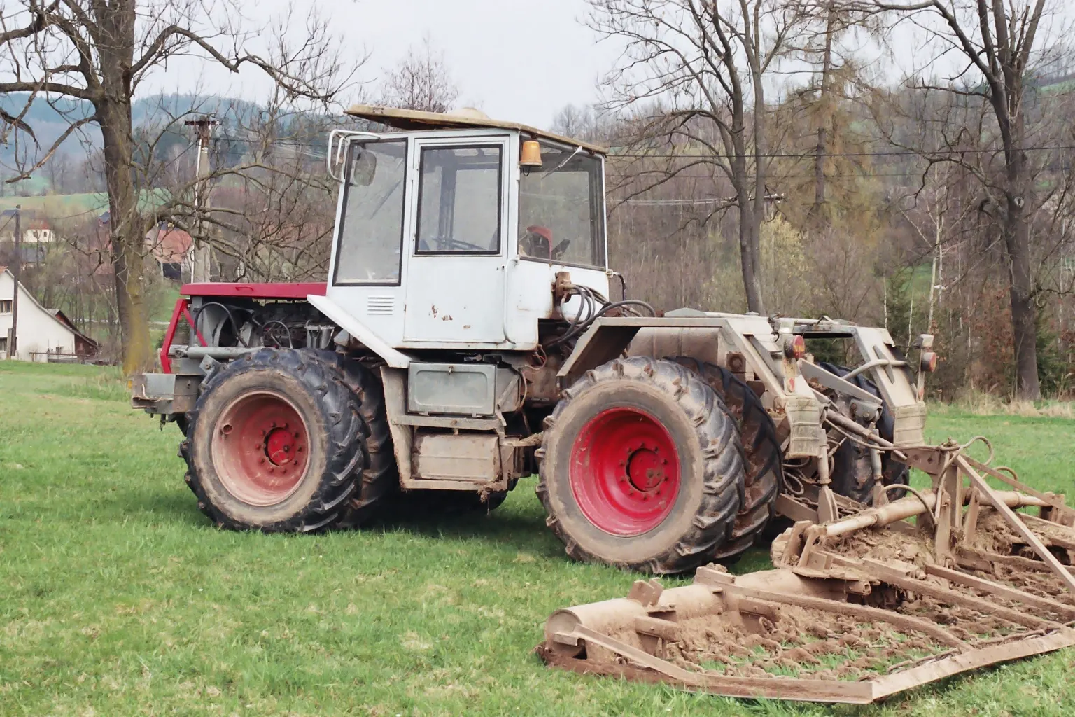 Tahač ŠT 180 z boku s bílou kabinou, červenou kapotou motoru, dvojmontáží kol a připojeným nářadím na louce