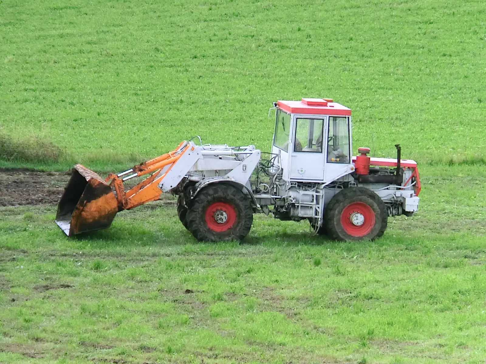 Bílý ŠT 180 N s červenou kapotou a oranžovým nakladačem z boku na louce