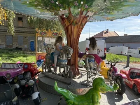 Malý kolotoč s dřevěnými koni a plastovými motorkami pod barevným stromem, dvě dívky na koních